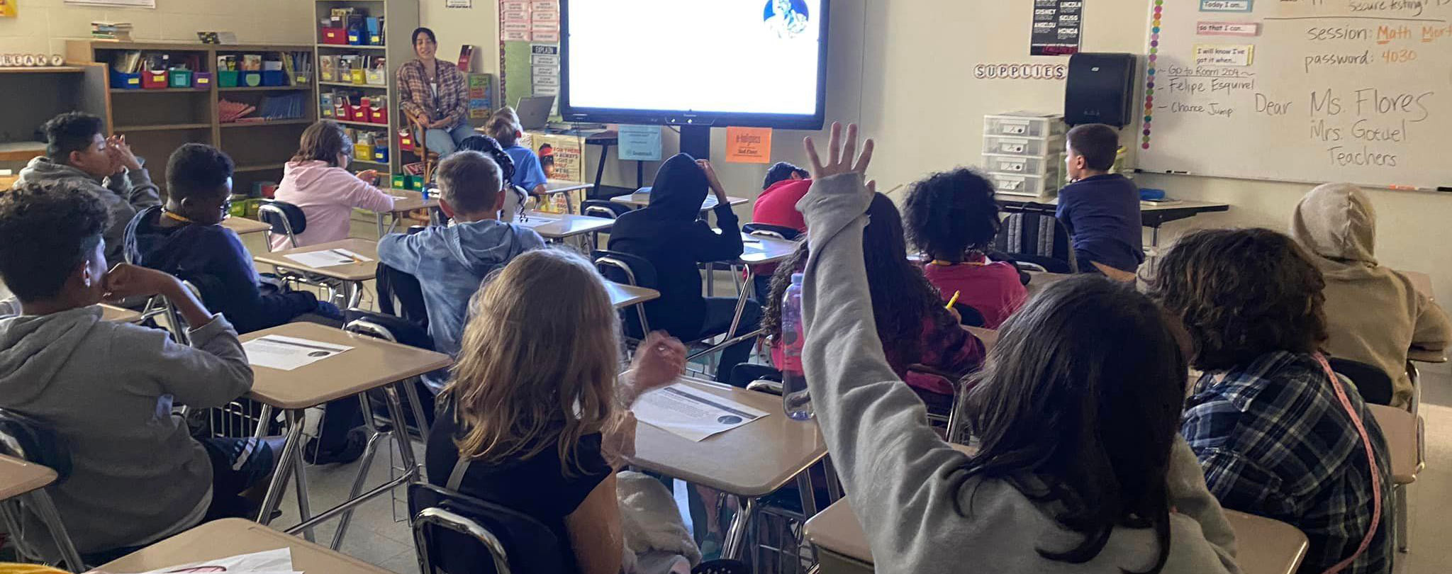 A wide shot of a classroom filled with students sitting at their desks, listening to their teacher. A student in the foreground is raising her hand.