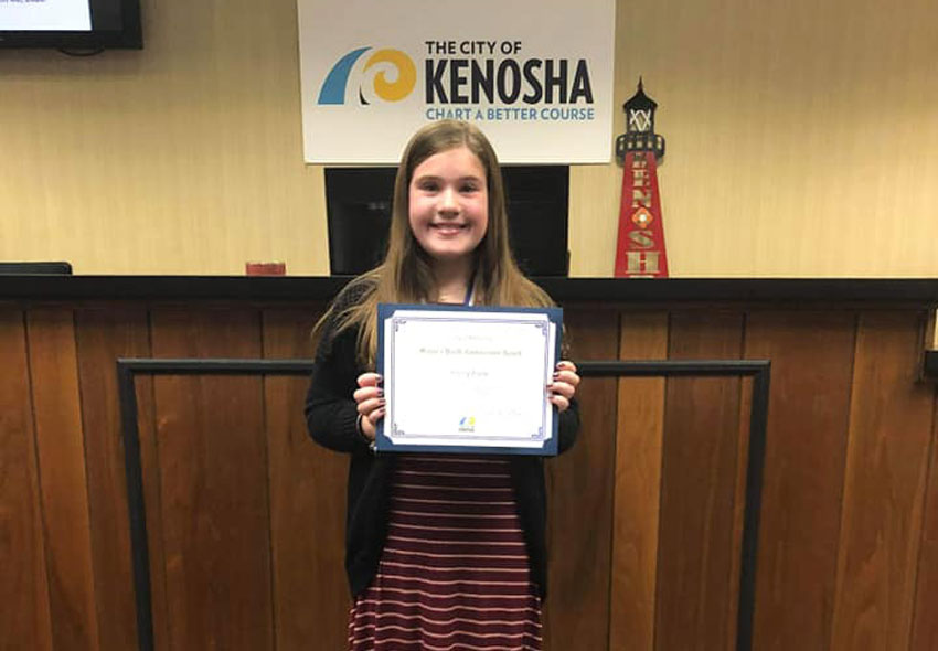 Emily holds her certificate in front of a city of Kenosha sign and model light house.