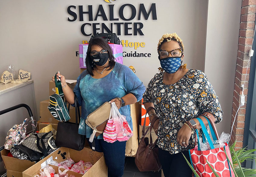 Two women holding donated purses in front of a Shalom Center sign.