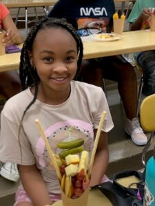 A girl smiles next to her charcuterie featuring two pickles.