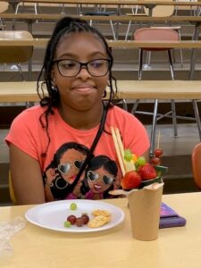 A girl wearing glasses smiles next to her charcuterie featuring two strawberries on top.