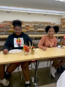 Two girls sitting at a table wearing glasses display their charcuterie creations.