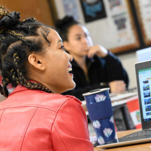 A woman wearing a red leather jacket, sitting next to a laptop smiles in class.