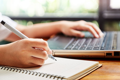 One hand on a laptop and another writing in a notebook.