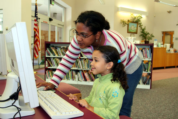 Second grade student working on a computer with help from a teacher.