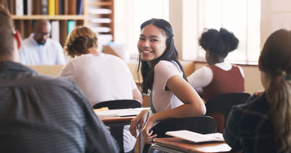 A student sitting at a desk among other students. She is facing towards the camera and smiling.