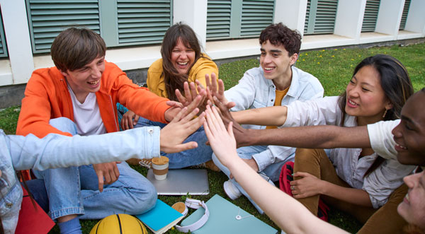 Five high school students sitting outside in a circle. Their hands are joined in the circle for a group high five.