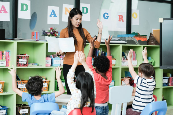A teacher reading a book to students who are raising their hands.